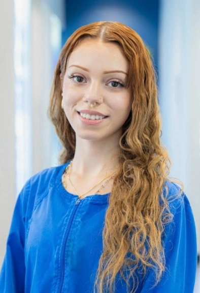 Young woman smiling at camera wearing dark shirt, blue dental office background with shelving.